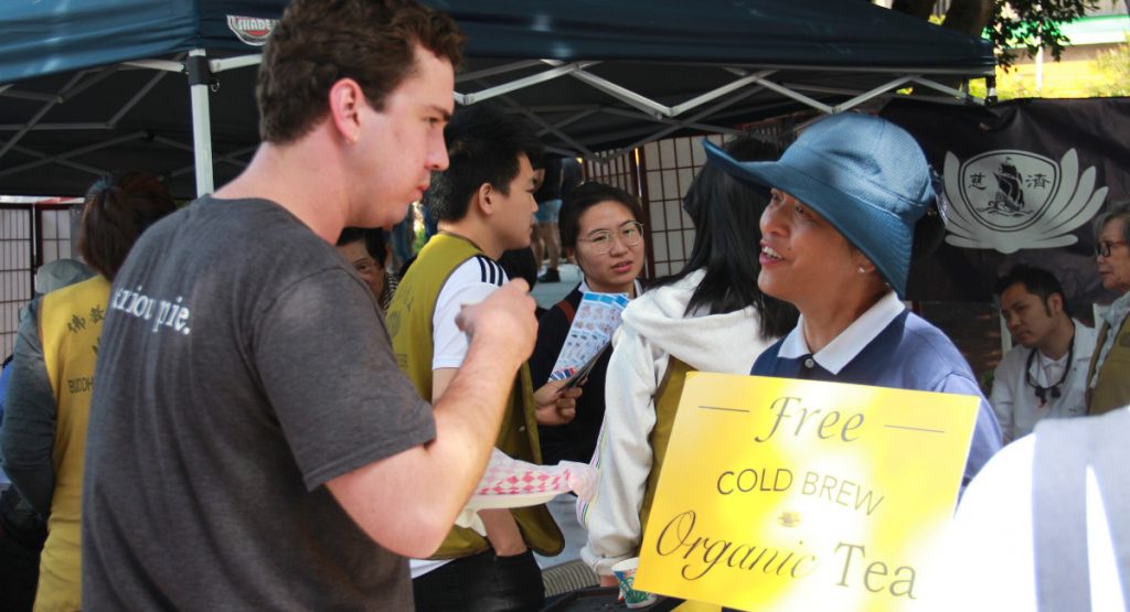 志工李淑娥手拿著自製的黃色免費有機冷泡茶招牌及環保毛毯，向遊客奉茶病並介紹慈濟。攝影／應仲銘