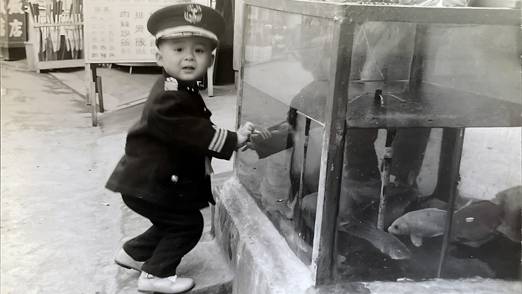 年幼的鄧博仁身穿小制服，在餐廳魚缸前留影。童真的神情定格成溫馨的時代記憶。圖片來源／鄧博仁