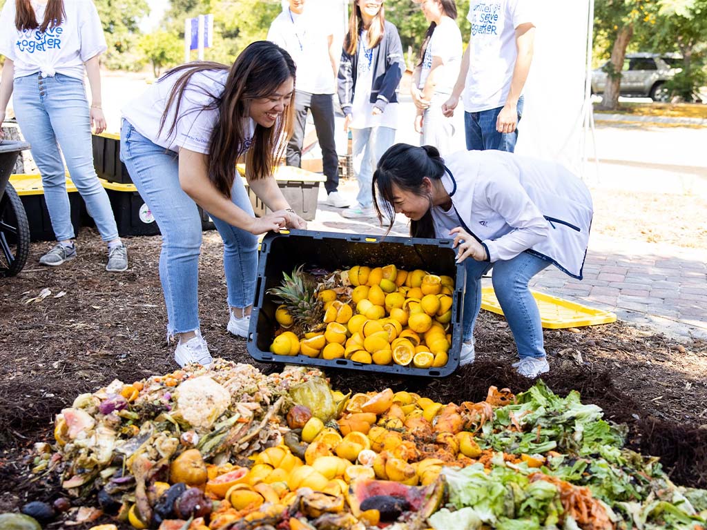 Volunteers adding food scraps to a large composting pile outdoors.