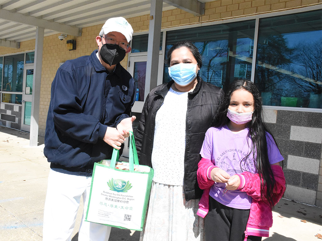 Volunteer hands a bag of supplies to a masked woman and girl outdoors.