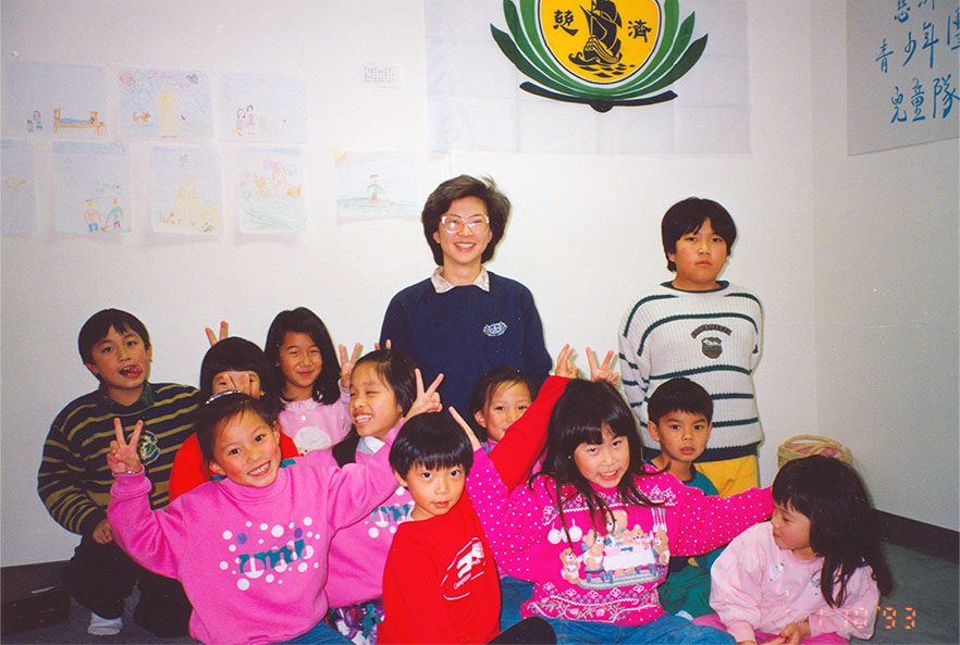 A woman in glasses poses with eleven happy children. The group gathers in a corner with drawings.