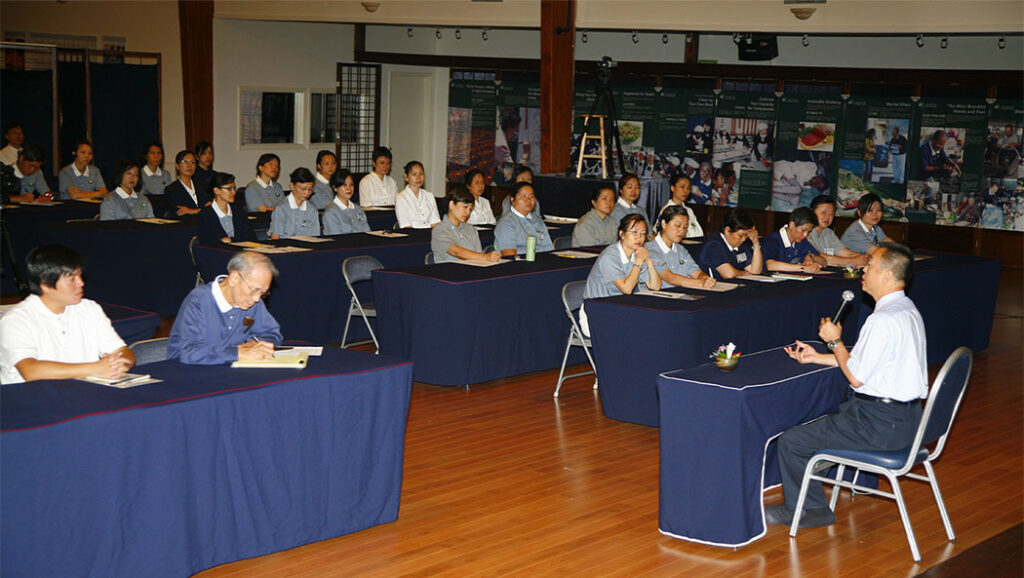 A group of Tzu Chi volunteers sits at tables in rows, listening to a man speaking.