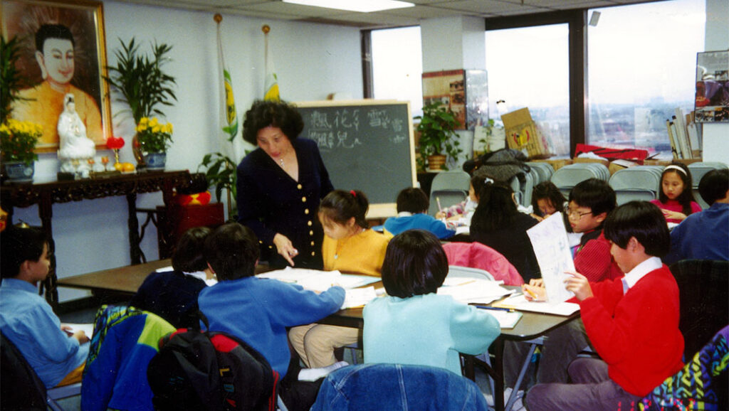 An adult Tzu Chi volunteer stands behind a table in a classroom teaching a group of about ten children who are sitting around a table with notebooks.